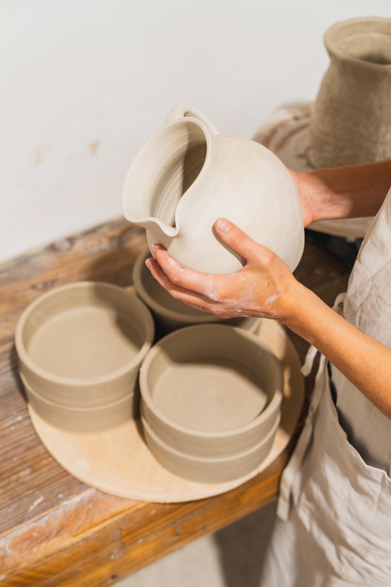 A potter holding an unfired clay jug above freshly thrown bowls